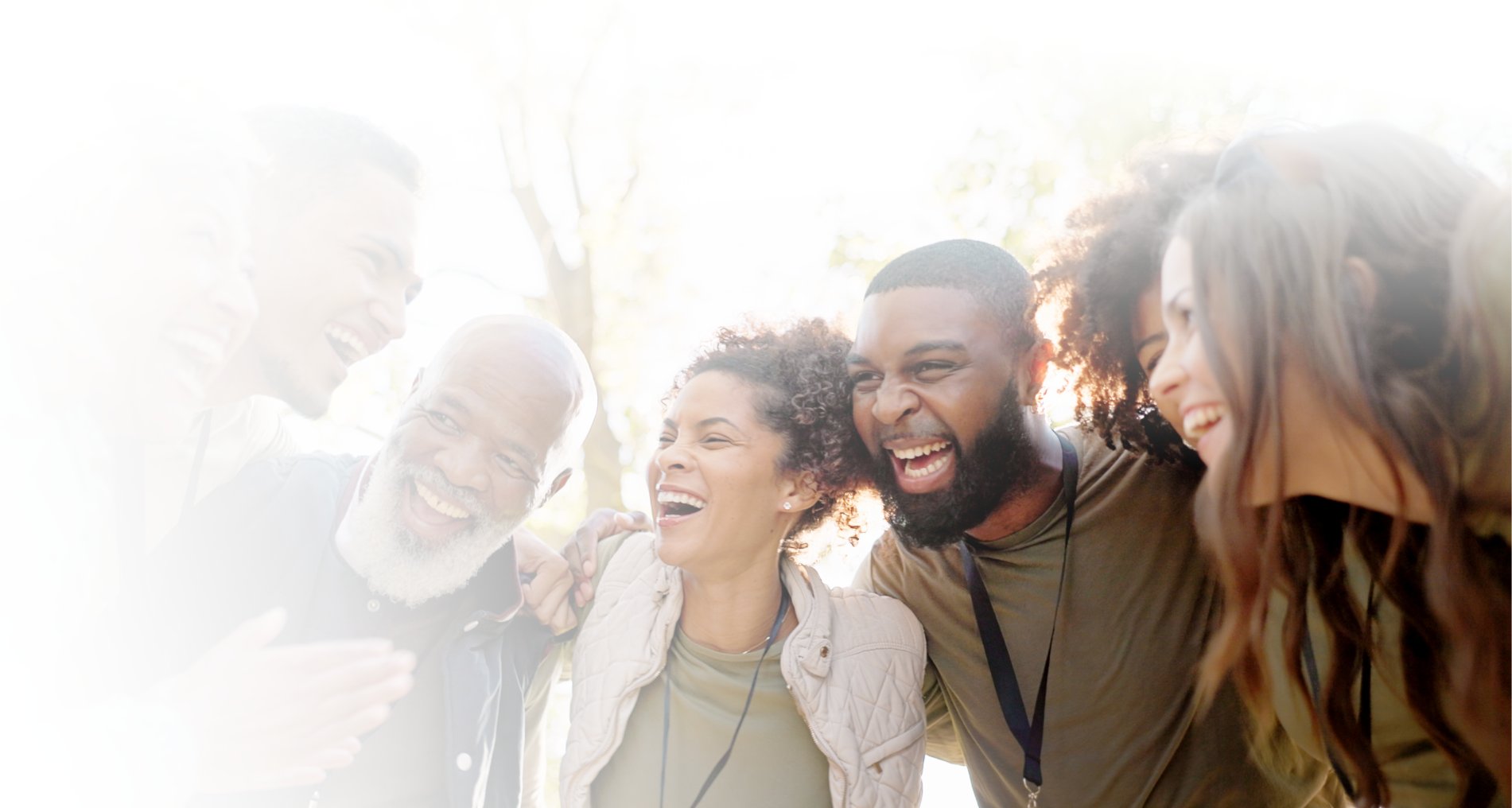 A group of six people stand close together outdoors, smiling and laughing, with sunlight in the background.