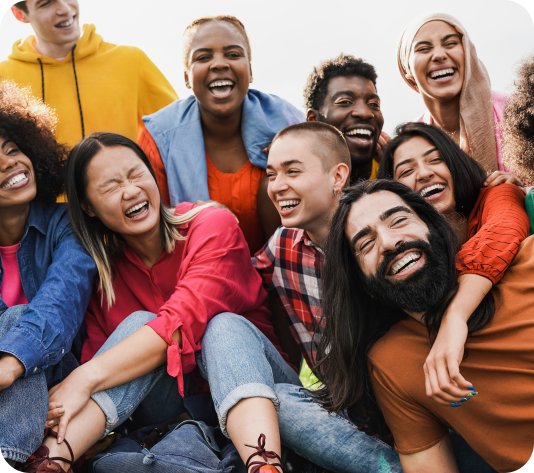 A diverse group of young adults sit closely together outdoors, smiling and laughing.