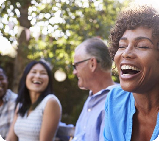 Four adults sit outdoors, smiling and laughing together, with trees and sunlight in the background.